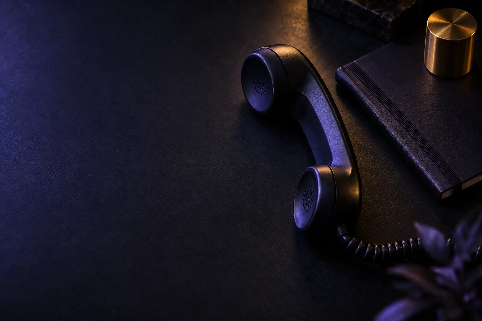 Auricular de teléfono negro sobre una mesa oscura junto a un cuaderno cerrado, iluminado con luz lateral violeta y un reflejo dorado.